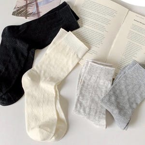 May include: A selection of socks in black, white, and grey, displayed on a table with an open book. The socks feature a textured, open-knit pattern. The socks are in shades of grey, white, and black.