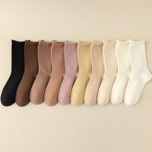 May include: A selection of ten pairs of crew socks in a range of neutral tones, such as black, brown, beige, and white. The socks are displayed in a line, highlighting their diverse colours and textures. The socks appear to be made of a soft material.