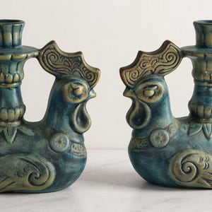 May include: Two teal and gold ceramic rooster candle holders. Each candle holder is shaped like a rooster with a handle and a candle cup. The rooster's comb and details are gold, and the body is teal.