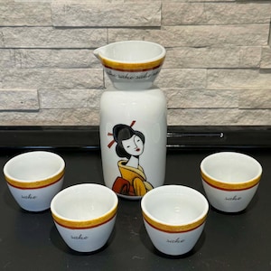 May include: A white sake set featuring a geisha design. The set includes a carafe, a small pitcher, and four sake cups. The cups and pitcher have a gold band around the rim, and the cups are labeled "sake".