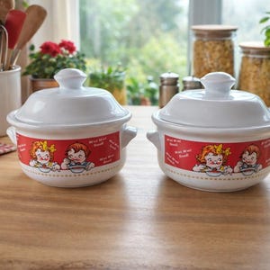 Peut inclure: Deux bols à soupe en céramique blanche avec couvercles. Chaque bol a une bande rouge avec une illustration de dessins animés d'enfants mangeant de la soupe. Les bols ont de petites poignées.