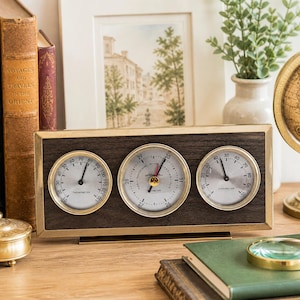 May include: A wooden weather station with three gauges in a brass frame. The gauges show temperature, humidity, and pressure. Other items include books, a globe, and a magnifying glass. The measurements are in Celsius.