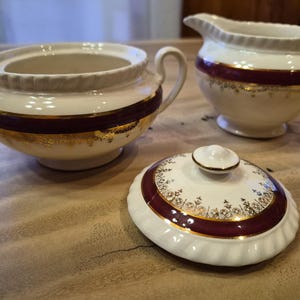May include: A cream-coloured ceramic sugar bowl, creamer, and lid set. Each piece features a burgundy band with gold filigree and trim. The sugar bowl has two handles, and the creamer has a spout.