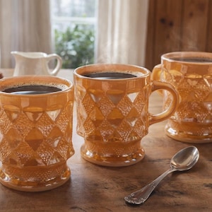 Peut inclure: Trois tasses à café ambrées remplies de liquide foncé, peut-être du café, sont posées sur une table en bois. Les tasses ont un motif de losanges texturés et sont accompagnées d'une cuillère et de muffins. De la vapeur s'élève des tasses.