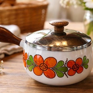 May include: White enamel saucepan with a stainless steel lid and wooden handle and knob. The pot is decorated with orange and green floral designs. A vintage kitchenware item.