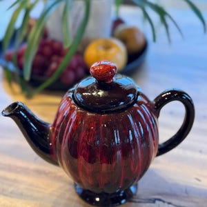 Peut inclure: Théière en céramique avec un design côtelé en forme de citrouille. La théière est principalement rouge foncé avec des accents noirs sur le bec, la poignée et le couvercle. Le couvercle a un petit bouton rouge. La théière est posée sur une surface en bois.