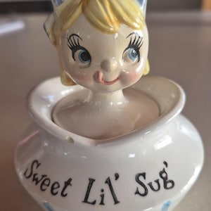 Peut inclure: Un sucrier en céramique avec un couvercle surmonté d'une figurine aux cheveux blonds portant un nœud à carreaux bleus et blancs. Le bol blanc a une forme arrondie et est décoré du texte noir Sweet Lil' Sug et de rayures bleues verticales.