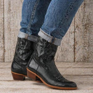 May include: Black leather cowboy boots with decorative stitching and a wooden heel. The boots are worn with blue jeans, cuffed at the ankles. The boots have a pointed toe and a low heel.