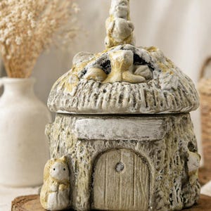Peut inclure: Une boîte à bijoux en céramique beige en forme de cottage. Le couvercle est orné d'une figurine d'écureuil. La maisonnette a une petite porte et un extérieur texturé. Elle repose sur une base en bois.
