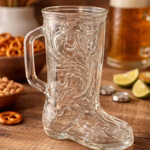 May include: Clear glass boot-shaped beer mug with an ornate floral design. The mug has a handle and is sitting on a wooden surface. Other items on the wooden surface include pretzels, beer, and lime slices.