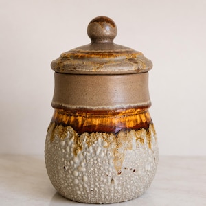 Peut inclure: Un pot en céramique avec un couvercle. Le pot est un mélange de marron, de beige et de blanc avec une surface texturée et tachetée. Le couvercle a un bouton arrondi et un bord décoratif. Le design du pot présente un dégradé de couleurs.