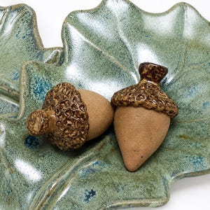 May include: Two ceramic acorns with smooth tan bodies and textured, glossy brown caps resting on a pair of overlapping, leaf-shaped ceramic dishes. The dishes feature a mottled, glossy green glaze with visible veining and irregular edges.