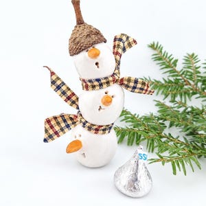 Op de afbeelding: Een witte keramieken sneeuwpopfiguur met drie gestapelde bollen, oranje wortelvormige neuzen en een bruine eikelkap als hoed. De sneeuwpop draagt een geruite stoffen sjaal met blauwe, rode en gele patronen. Een zilverkleurig verpakt chocolaatje ligt ernaast.
