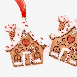 May include: Two gingerbread house ornaments with red ribbon hangers. The houses are decorated with white frosting, red sprinkles, candy canes, and heart-shaped details. A small, silver-wrapped candy is also present.
