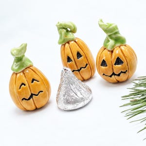 Op de afbeelding: Drie kleine keramische pompoen-lantaarns met glanzende oranje lichamen en lichtgroene stelen, elk voorzien van een handgeschilderd zwart uitgesneden gezicht. Een enkele in zilverfolie verpakte chocoladedruppel ligt in het midden van het geheel.