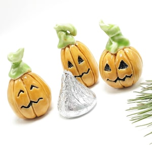 May include: Three ceramic jack-o'-lantern figurines with green stems and carved faces. A silver-wrapped chocolate candy sits in front of the pumpkins. The pumpkins are orange with black facial features.