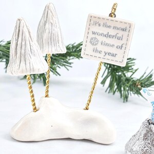 May include: A white ceramic base supports two textured white tree sculptures and a square white sign with the text It's the most wonderful time of the year and a snowflake icon. The pieces are connected by thin, twisted gold-colored metal rods.