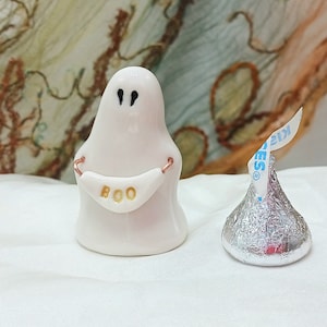 May include: A small white ceramic ghost figurine with black painted eyes, holding a white banner with the gold text BOO, positioned next to a silver foil-wrapped chocolate candy with a white paper plume labeled Kisses in blue text.
