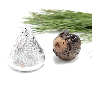 May include: A small, round, dark brown ceramic hedgehog figurine with a smooth face and textured body, positioned next to a single chocolate candy wrapped in shiny silver foil. The hedgehog has two small ears and a simple, carved facial expression.