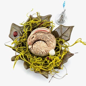 May include: A ceramic sleeping bunny figurine nestled in a leaf-shaped dish with green moss. The bunny is light brown with pink ears and a white tail. A small red ladybug is on the dish. A silver-wrapped chocolate kiss is in the background.