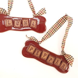 May include: Two glossy, maroon-colored dog bone ornaments with names spelled out in individual letter tiles. One says "LUNA" and the other says "PEPPER". Each bone has a plaid ribbon for hanging.