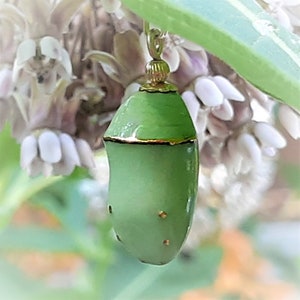 Op de afbeelding: Een close-up van een groene cocon met gouden accenten, hangend aan een delicate gouden ketting. De cocon heeft kleine gouden stippen en een gouden band aan de bovenkant. Zachte achtergrond van witte bloemen en groene bladeren.