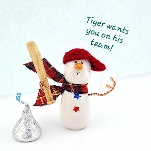 May include: A whimsical snowman figurine with a red knit cap and scarf, holding a wooden baseball bat and a baseball. The snowman has a carrot nose and star accents. A wrapped chocolate candy is in the foreground. Text reads: "Tiger wants you on his team!"