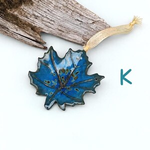 Op de afbeelding: Een glanzend blauw keramieken esdoornbladornament met donkerbruine nerven en spikkels. Het blad heeft een lichtbeige lint dat door een klein gaatje aan de bovenkant is gestrikt om op te hangen. De letter K staat in het groenblauw rechts.