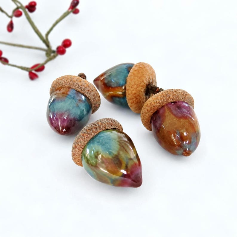 May include: Three decorative ceramic acorns featuring glossy, multi-coloured glazes in shades of blue, brown, and magenta. Each acorn has a textured, tan-coloured cap with a realistic appearance, arranged on a plain white surface.