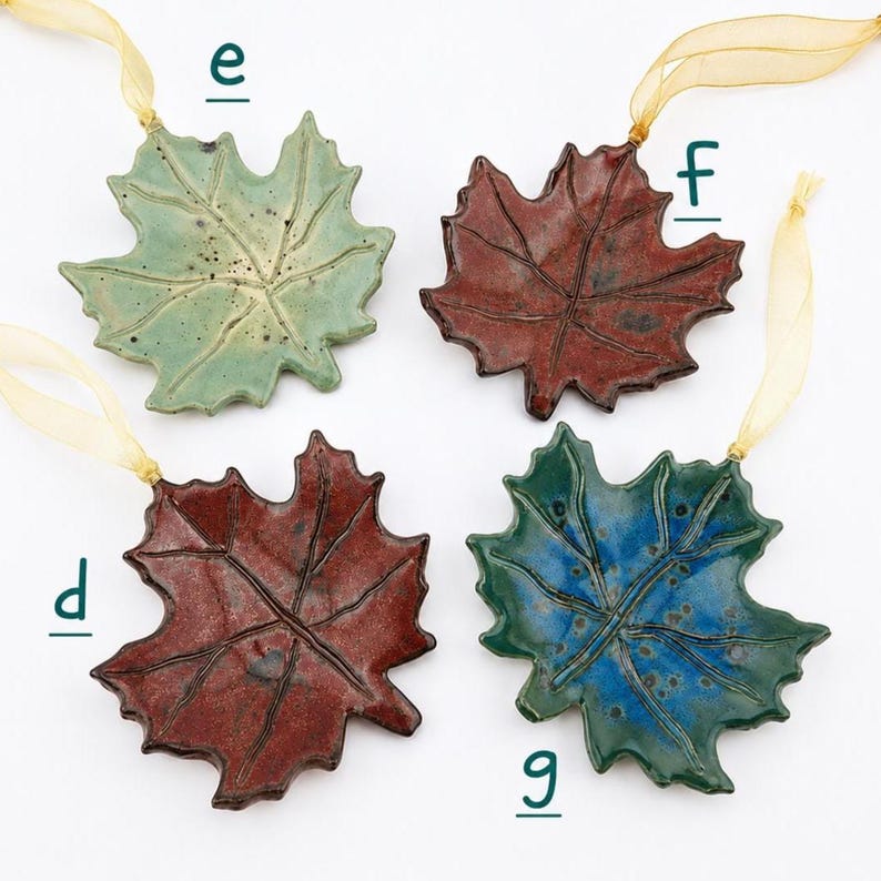 Op de afbeelding: Vier keramische esdoornbladhangers met glanzende glazuren in lichtgroen, donkerrood en blauw, elk voorzien van een gouden lintlus om op te hangen. De hangers zijn gemarkeerd met de letters e, f, d en g in een donkergroen lettertype.