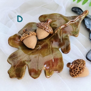 May include: A ceramic oak leaf dish with a glossy green and brown glaze, featuring two attached acorn sculptures with textured brown caps and smooth tan bodies, plus one separate matching acorn sculpture placed to the side, marked with the letter D.