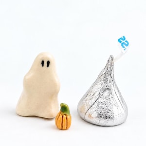 May include: A small off-white ceramic ghost figurine with two black painted eyes, a tiny orange ceramic pumpkin with a green stem, and a silver foil-wrapped chocolate candy with a small blue and white paper tag protruding from the top.