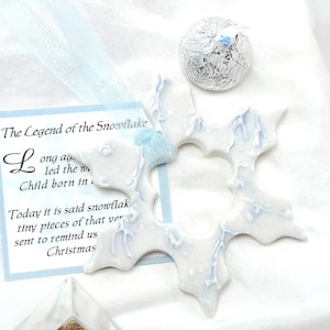 May include: White snowflake ornament with light blue accents and a light blue ribbon. The ornament is next to a small, silver-wrapped sphere and a card with the text "The Legend of the Snowflake".
