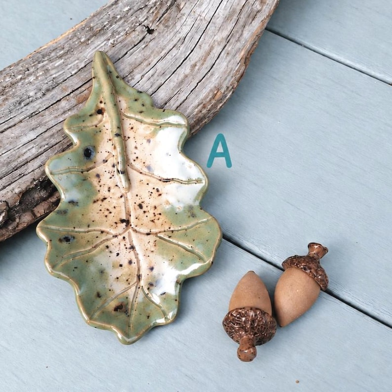 May include: A ceramic leaf-shaped dish with a glossy green and cream glaze featuring dark brown speckles, marked with the letter A. Beside the dish are two small ceramic acorn figurines with textured brown caps and smooth tan bodies.