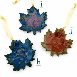 Op de afbeelding: Drie keramische esdoornbladornamenten met glanzende afwerking en linten. Ornament h is diepblauw met groene spikkels, i is blauw met een paars centrum, en j is een mix van bruin en groen. Elk blad heeft gedetailleerde bladnerven.