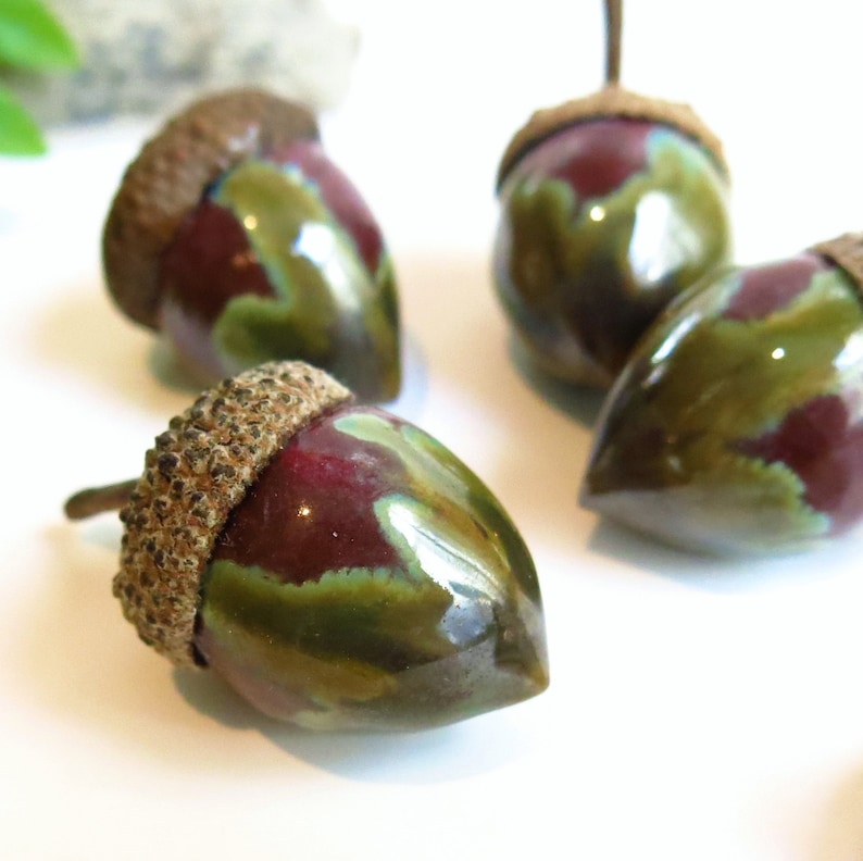 May include: A set of three ceramic acorn ornaments featuring a glossy finish with blended deep burgundy and olive green colours. Each acorn has a textured, brown, realistic cap and a smooth, pointed nut body, arranged in a cluster on a white surface.