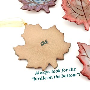 Op de afbeelding: Een verzameling keramische esdoornbladornamenten met glanzende rode en blauwe glazuren. E&eacute;n ornament is omgedraaid om een matte, lichtbruine onderkant te tonen met een kleine vogelstempel en de tekst "Always look for the birdie on the bottom"!