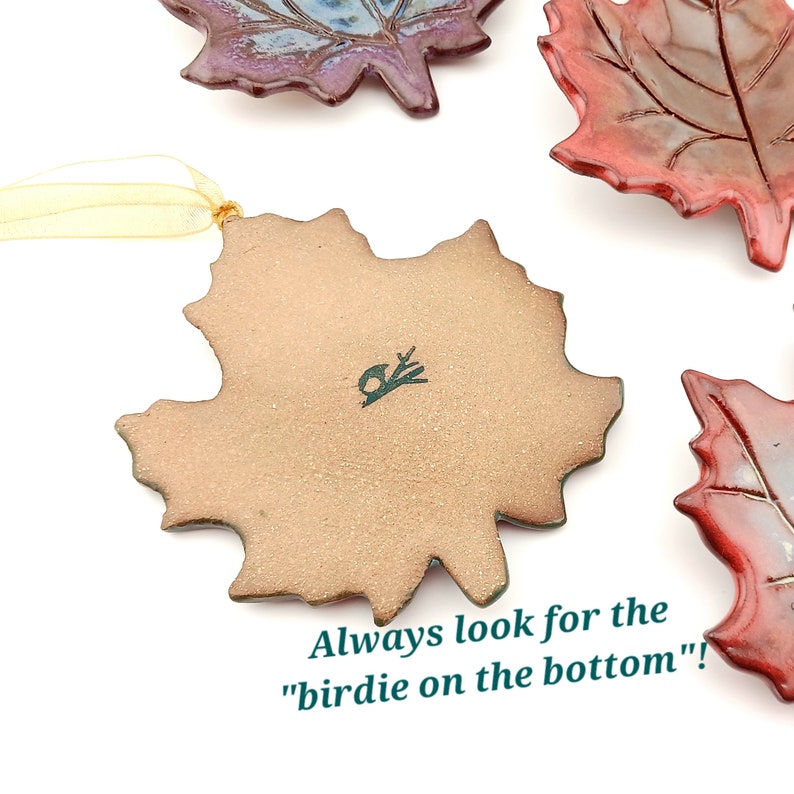 Op de afbeelding: Een verzameling keramische esdoornbladornamenten met glanzende rode en blauwe glazuren. E&eacute;n ornament is omgedraaid om een matte, lichtbruine onderkant te tonen met een kleine vogelstempel en de tekst "Always look for the birdie on the bottom"!