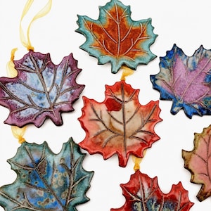 Op de afbeelding: Een verzameling van zeven keramische esdoornbladornamenten in diverse kleuren, waaronder blauw, paars, rood en oranje. Elk blad heeft een glanzend glazuur en gedetailleerde nerven, sommige zijn voorzien van gele linten.
