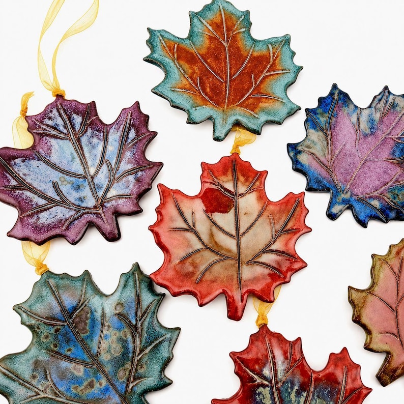 Op de afbeelding: Een verzameling van zeven keramische esdoornbladornamenten in diverse kleuren, waaronder blauw, paars, rood en oranje. Elk blad heeft een glanzend glazuur en gedetailleerde nerven, sommige zijn voorzien van gele linten.