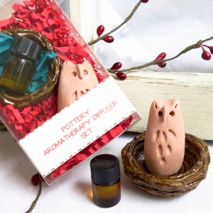 May include: A pottery aromatherapy diffuser set featuring a light brown owl figurine and a small brown bottle. The set includes a ceramic owl in a nest-like holder, a small glass bottle, and a clear box with red paper filler. The text on the box reads "Pottery Aromatherapy Diffuser Set."