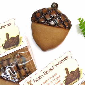 May include: A ceramic acorn bread warmer with a brown, textured top and a tan base. The acorn is on a white surface with a package of smaller acorn-shaped warmers and a tag that reads "Acorn Bread Warmer."