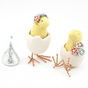 May include: Two yellow ceramic chicks with flower crowns, each sitting in a white ceramic eggshell. The chicks have copper wire legs and are on a white surface.