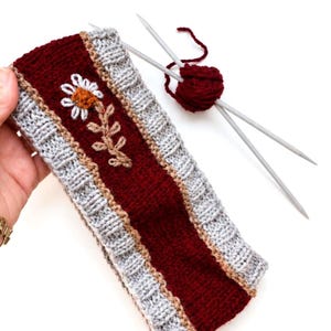 Op de afbeelding: Een gebreide hoofdband met een diepbordeauxrood middenpaneel met een wit en oranje madeliefjesborduursel, omrand door lichtgrijze geribbelde randen en dunne beige strepen, vergezeld van twee breinaalden en een bolletje bordeauxrood garen.