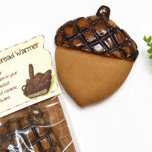 May include: A ceramic bread warmer shaped like an acorn with a glossy dark brown cap and a matte light brown base. The packaging includes a label with the text Bread Warmer and an illustration of bread in a basket, with another acorn warmer visible.