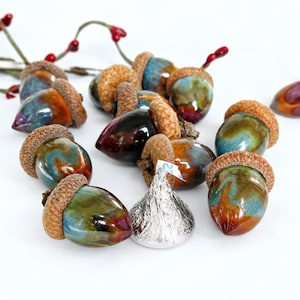 May include: A collection of ceramic acorns with glossy, multi-coloured glazes in shades of blue, brown, and red, each topped with a textured brown cap. A single silver foil-wrapped chocolate kiss sits among the acorns, which are arranged near red berries.