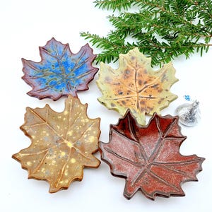 May include: Four ceramic leaf-shaped dishes in shades of blue, green, brown, and red. Each dish has detailed veining and a glossy finish. The dishes are arranged on a white surface with a sprig of greenery and a wrapped chocolate candy.