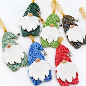 May include: Six ceramic gnome ornaments in green, light green, blue, red, and brown, each featuring a textured leaf pattern, a white clay beard, a small nose, and a silver bell attached to a gold ribbon loop for hanging on a tree or display.