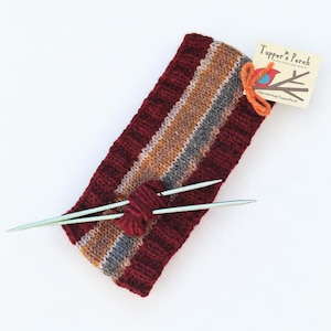 Op de afbeelding: Een gebreide hoofdband met een brede bordeauxrode geribbelde rand en een centraal streeppatroon in grijstinten, beige en bordeaux. Twee zilveren breinaalden zijn door een kleine wollen knoop gestoken. Een label vermeldt Tupper's Perch.