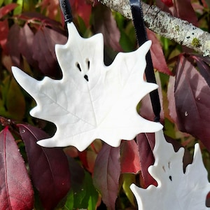 May include: A white ceramic maple leaf ornament featuring a ghost-like face with two small eye cutouts and a tiny mouth hole. The ornament is suspended by a thin black ribbon, showcasing a smooth finish and detailed leaf vein textures.
